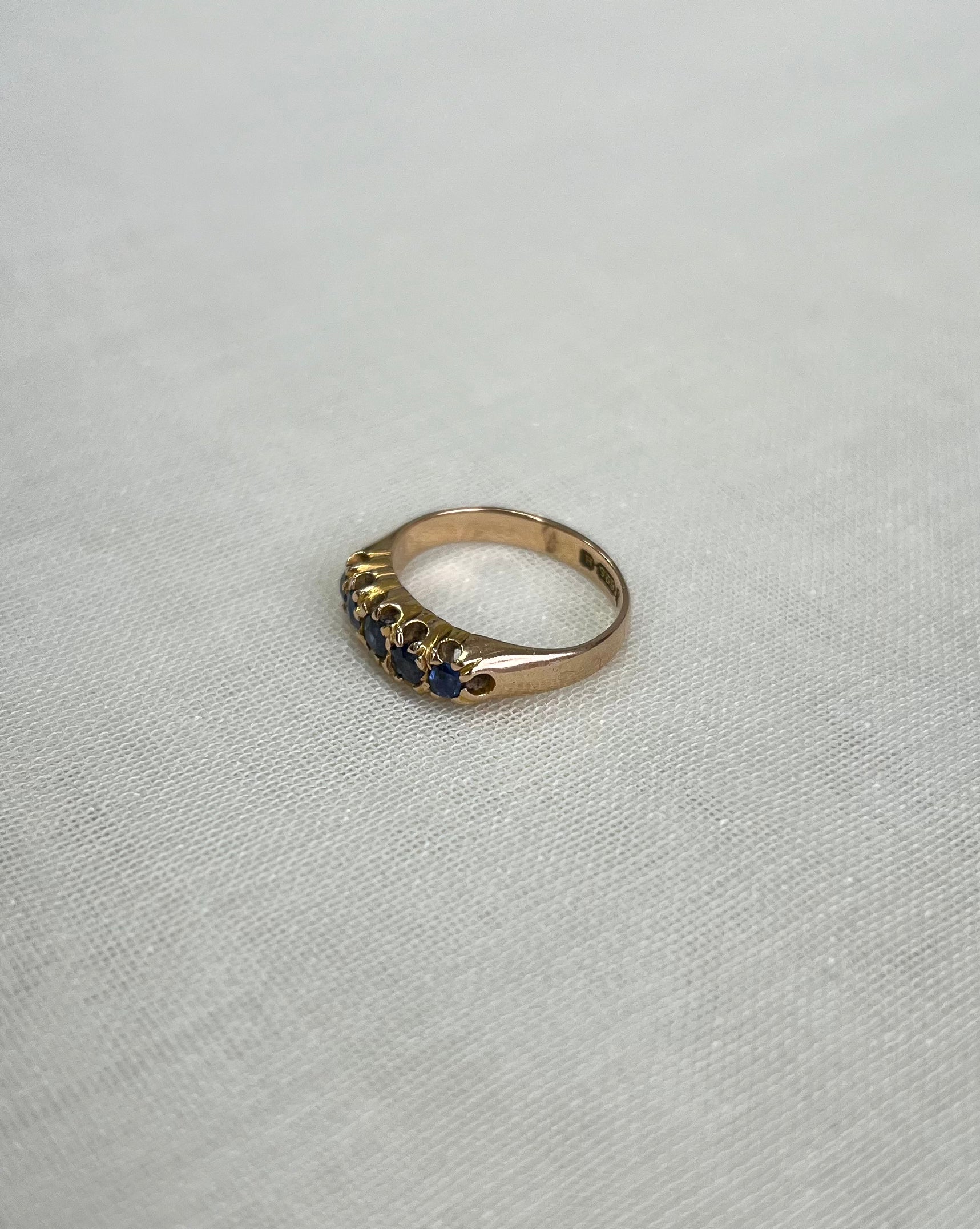 A gold ring with a row of small dark blue gemstones, photographed on a light cream coloured textured fabric background.