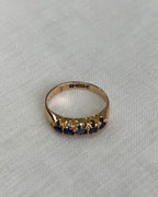 A yellow gold ring featuring a row of dark blue gemstones, photographed on a textured light beige fabric background.