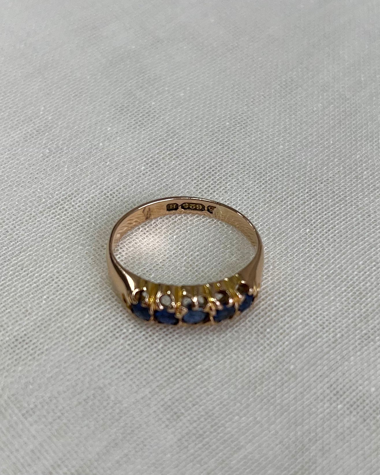 A yellow gold ring featuring a row of dark blue gemstones, photographed on a textured light beige fabric background.