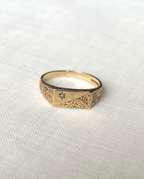 A gold rectangle ring set with diamond in starburst engraving and intricate pattern engraving, resting on white fabric background 