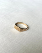 A gold signet ring with engraved geometric patterns and a small star detail set with diamond stone, resting on a textured white fabric background.