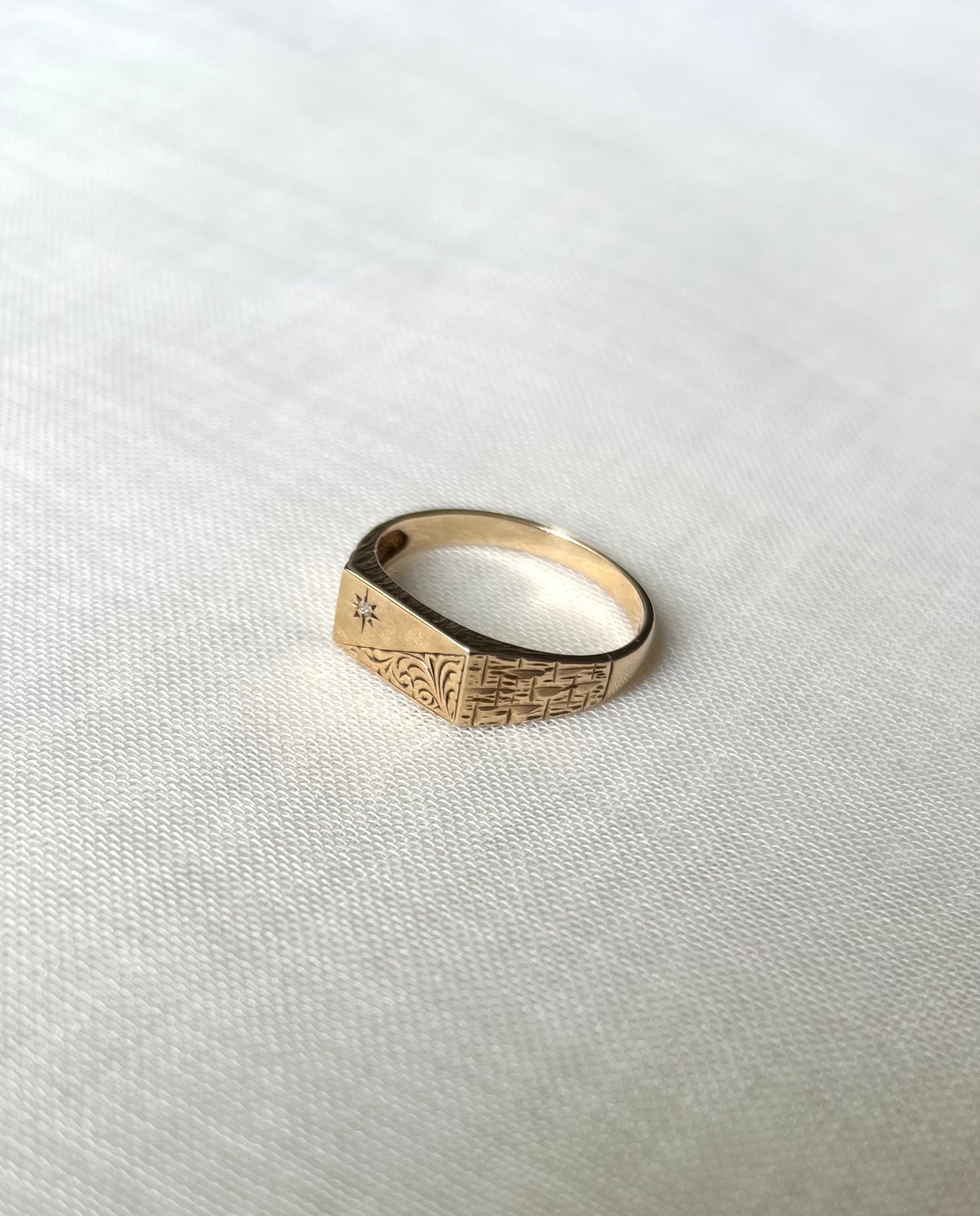 A gold signet ring with engraved geometric patterns and a small star detail set with diamond stone, resting on a textured white fabric background.