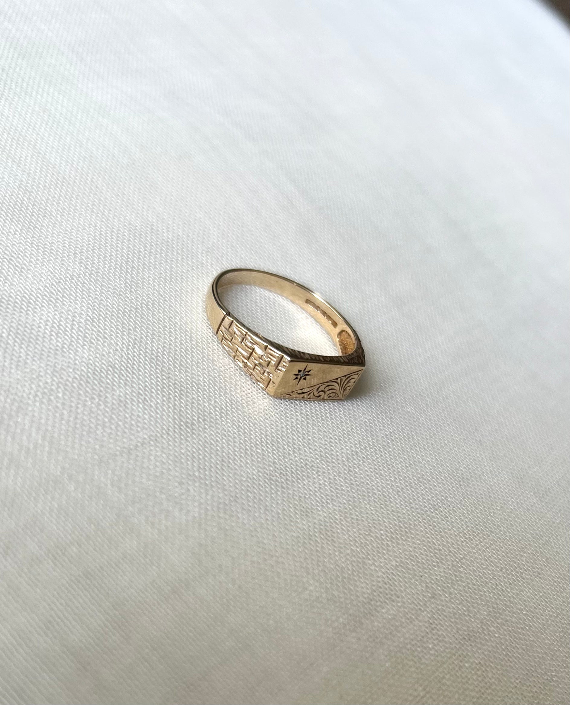 A gold signet ring with engraved geometric patterns and a small star detail set with diamond, resting on a textured white fabric background.