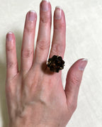 A close-up of a hand wearing a large gold ring with a dark brown smoky gemstone, against a light beige textured background.