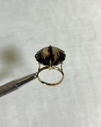 gold ring with large brown smokey quartz gemstone, held by silver tool above white background 