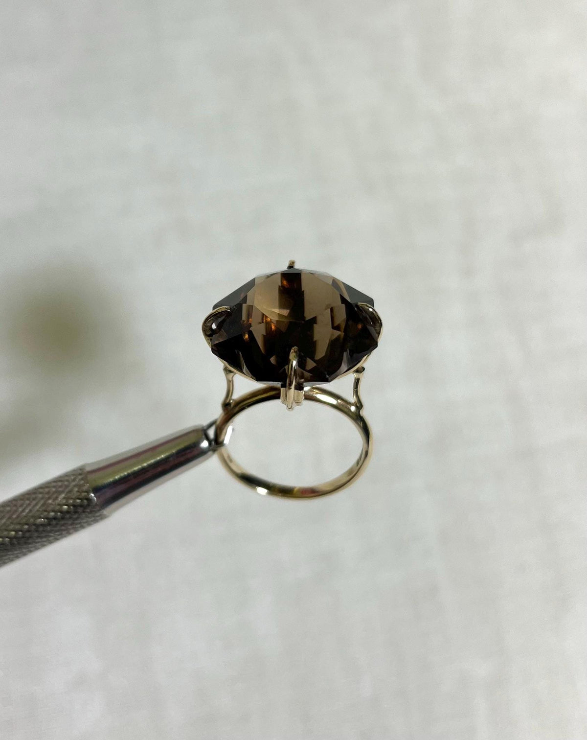 gold ring with large brown smokey quartz gemstone, held by silver tool above white background 
