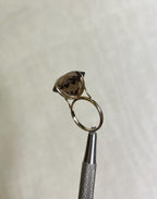 side view of large statement ring with brown smokey quartz gemstone, held by silver tool above white background