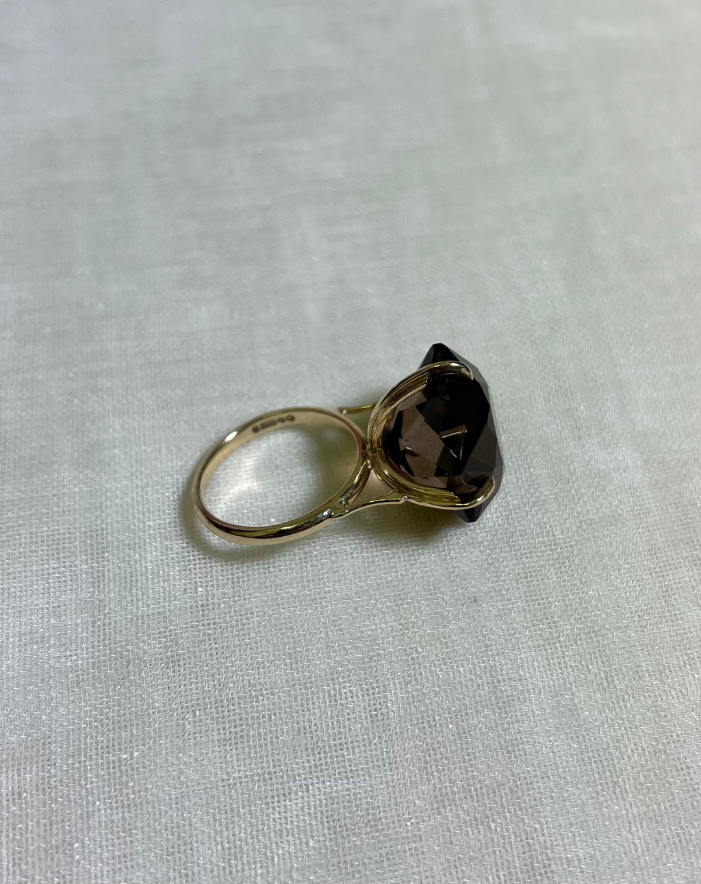 A gold ring with a large, faceted dark brown smoky gemstone, resting on a textured off-white fabric background.