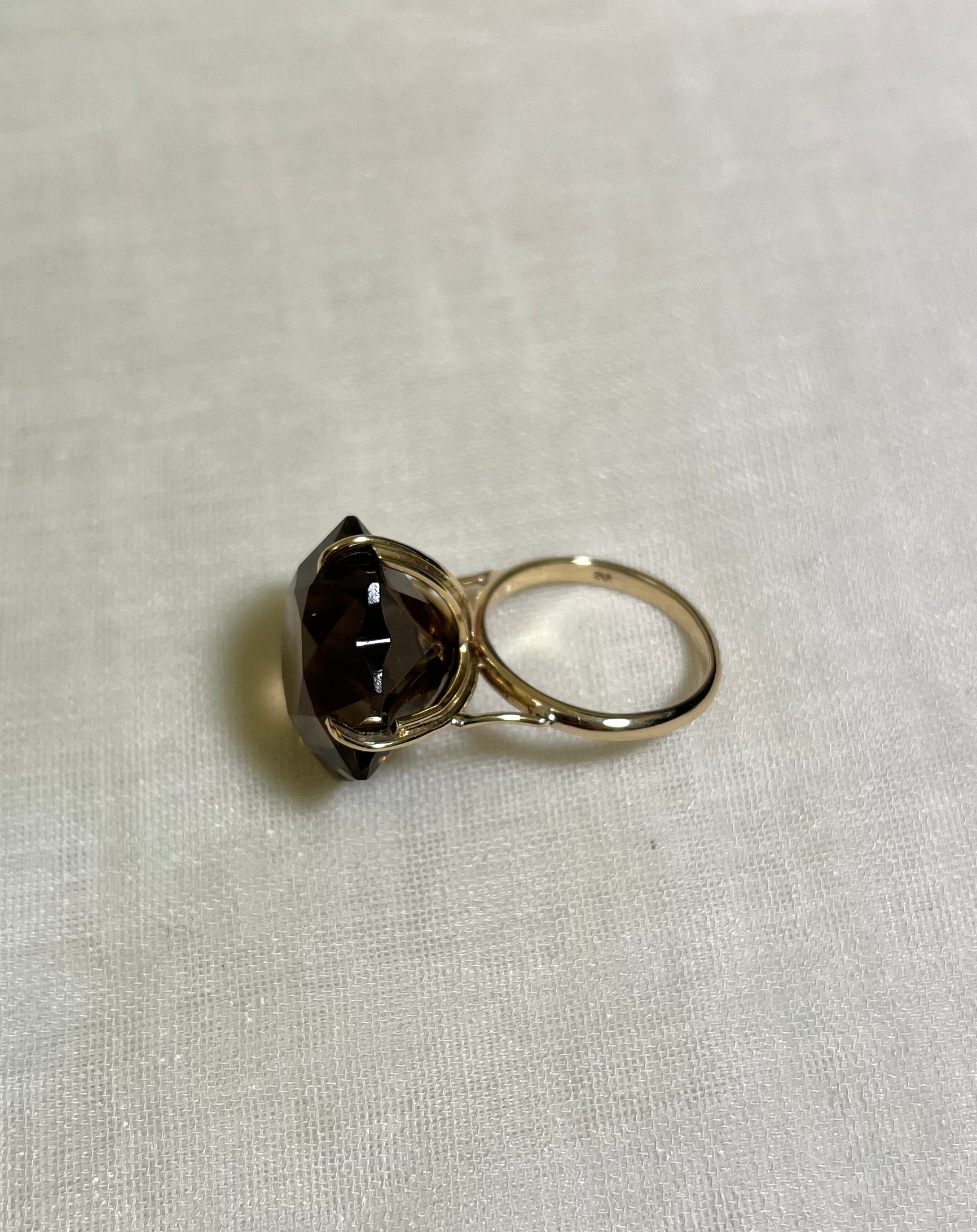 Side view of gold ring featuring a large, faceted dark brown smoky gemstone, photographed on a textured off-white fabric background.