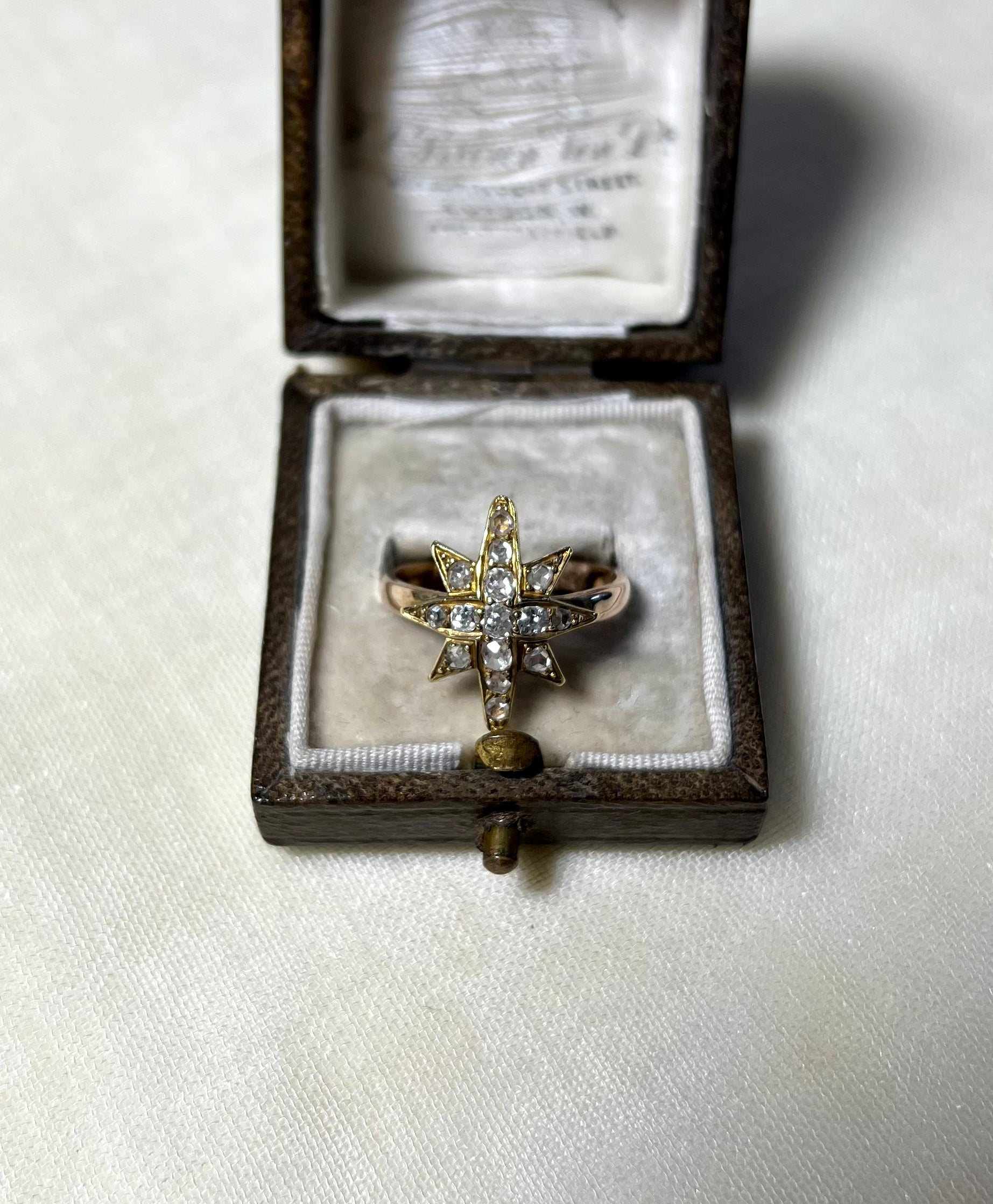 a gold star ring set with diamonds, displayed in brown box on white fabric background