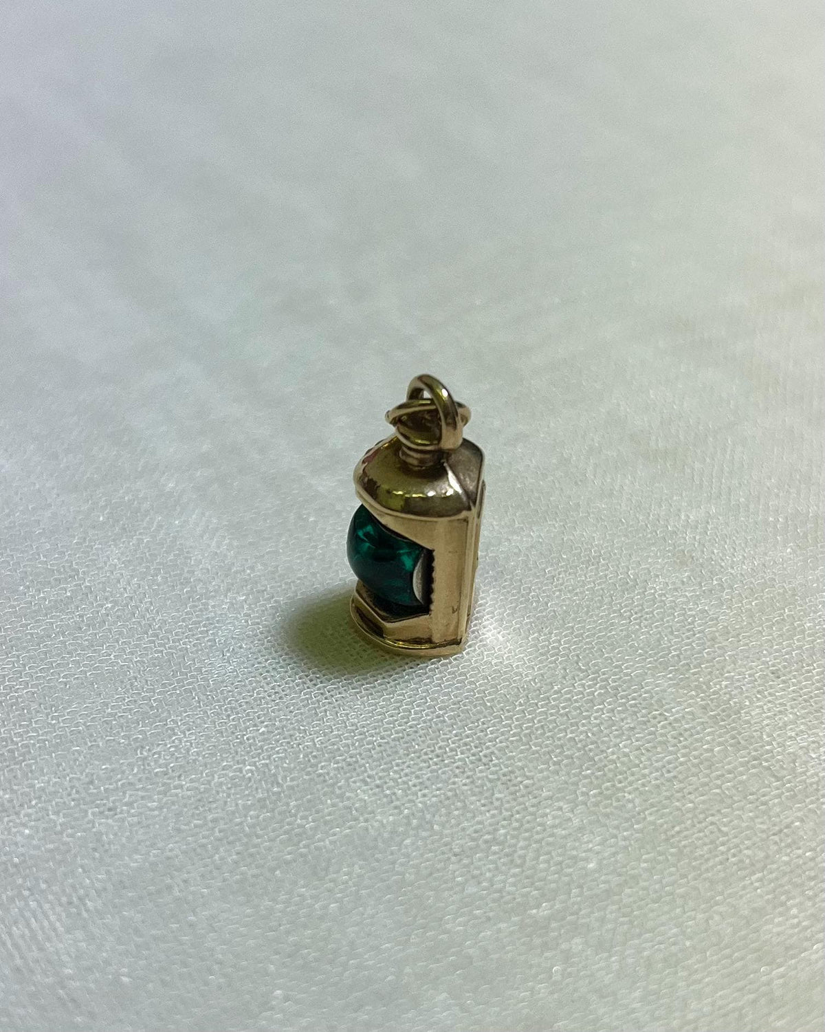 Gold pendant with a green centre in shape of ship lantern on a white fabric background
