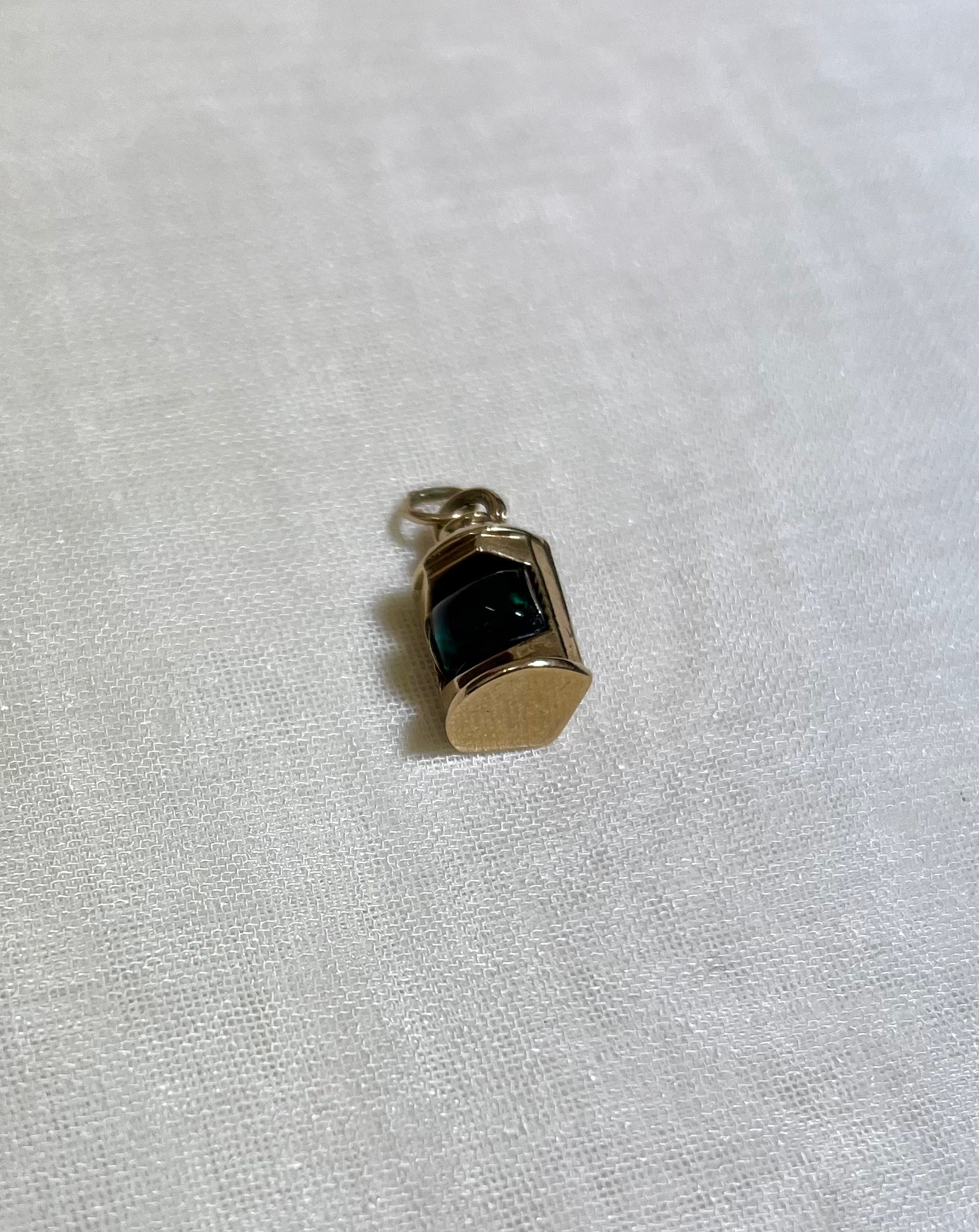 gold pendant in shape of ship lantern with green centre, resting on white fabric background 