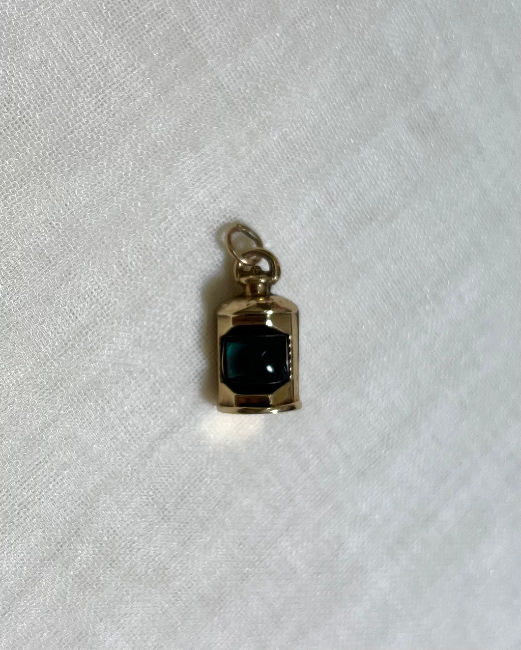 A small gold pendant charm with a dark green centre, resting on a textured white fabric background.