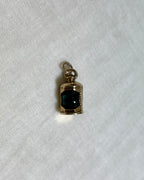 A small gold pendant charm with a dark green centre, resting on a textured white fabric background.
