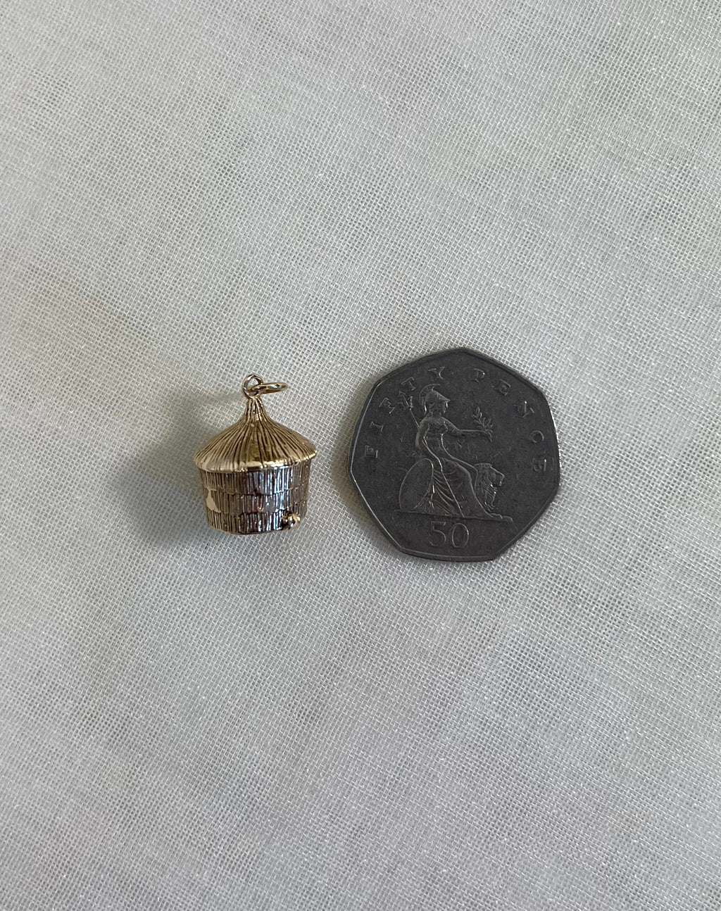 small straw hut gold charm nest to a 50p coin for size guide on light fabric background