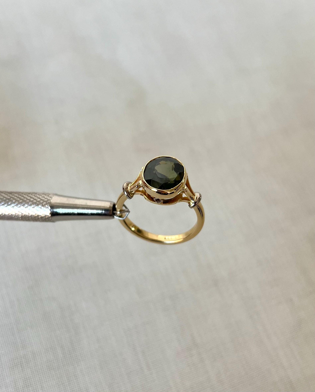 gold ring with dark green gemstone, held by silver tool above white background 
