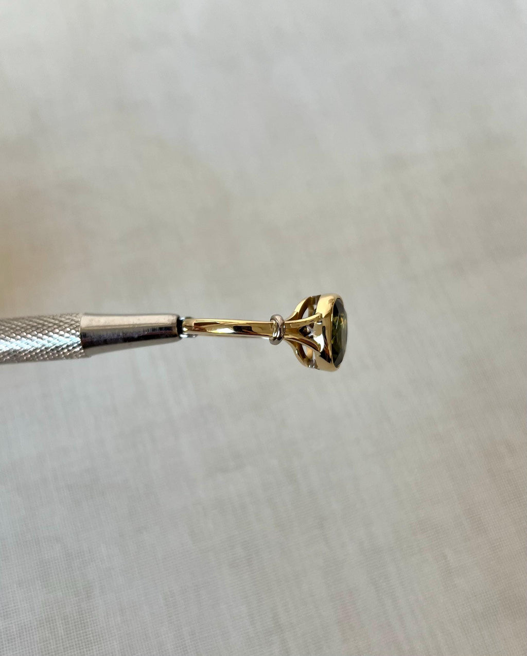 side view of gold ring with green gemstone held by silver tool above white background 