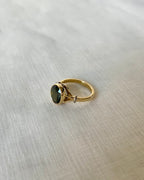 A gold ring with an oval dark green gemstone, resting on a textured beige fabric background.