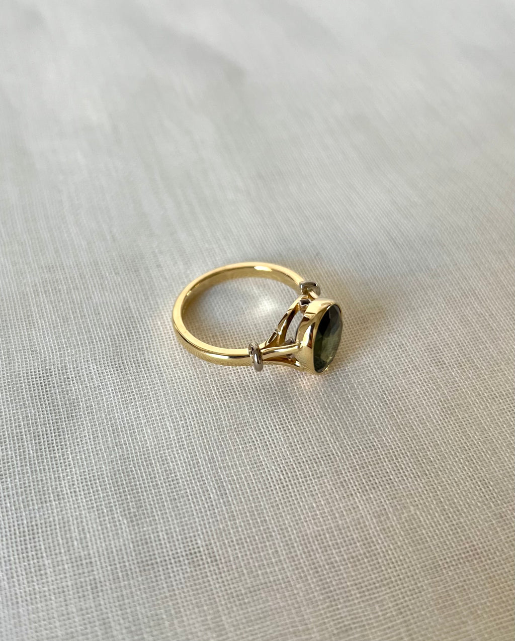 A gold ring with a dark green oval gemstone, resting on a textured light beige fabric background.