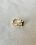 A gold ring with a dark green oval gemstone, resting on a textured light beige fabric background.