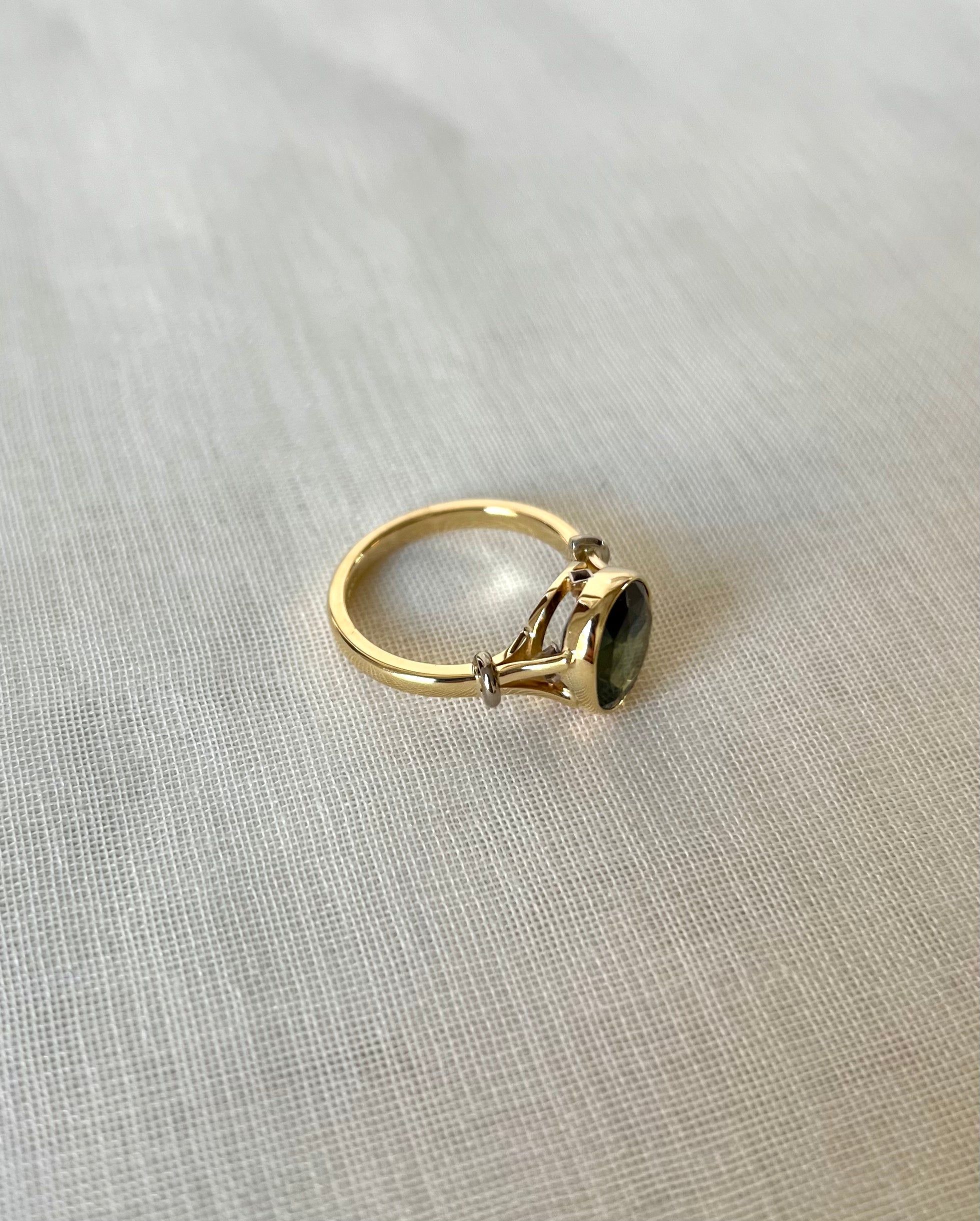 A gold ring with a dark green oval gemstone, resting on a textured light beige fabric background.
