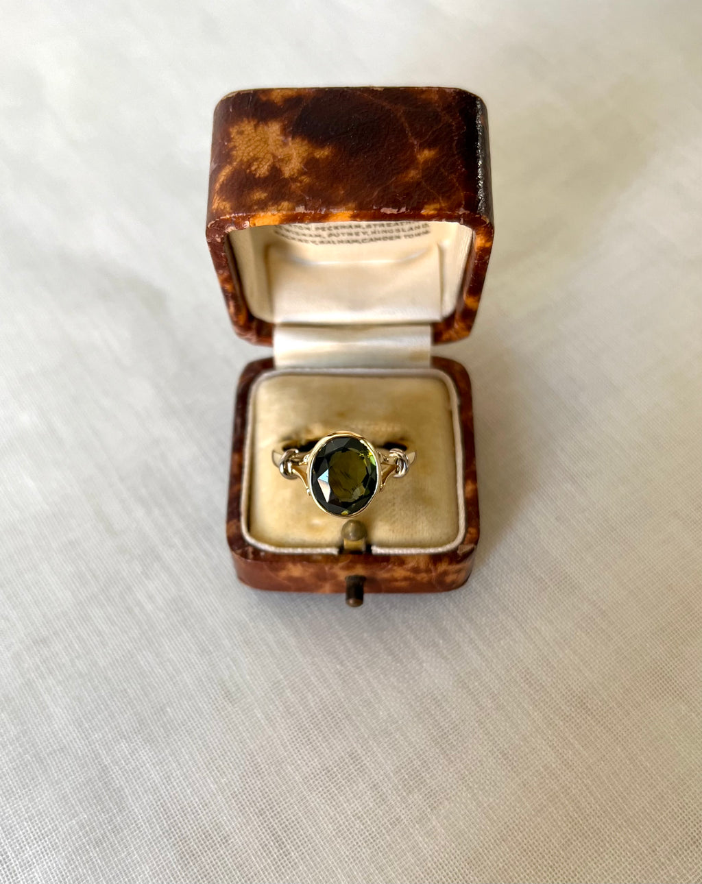 a gold ring with oval green gemstone in a brown fleck vintage ring box, resting on light beige fabric background 