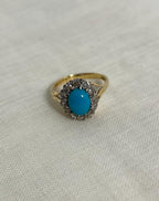 Gold ring with a turquoise stone surrounded by diamonds on a light fabric background