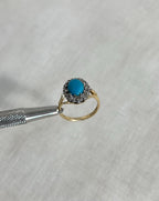 Gold ring with a turquoise gemstone and diamonds held by tweezers on a light fabric background