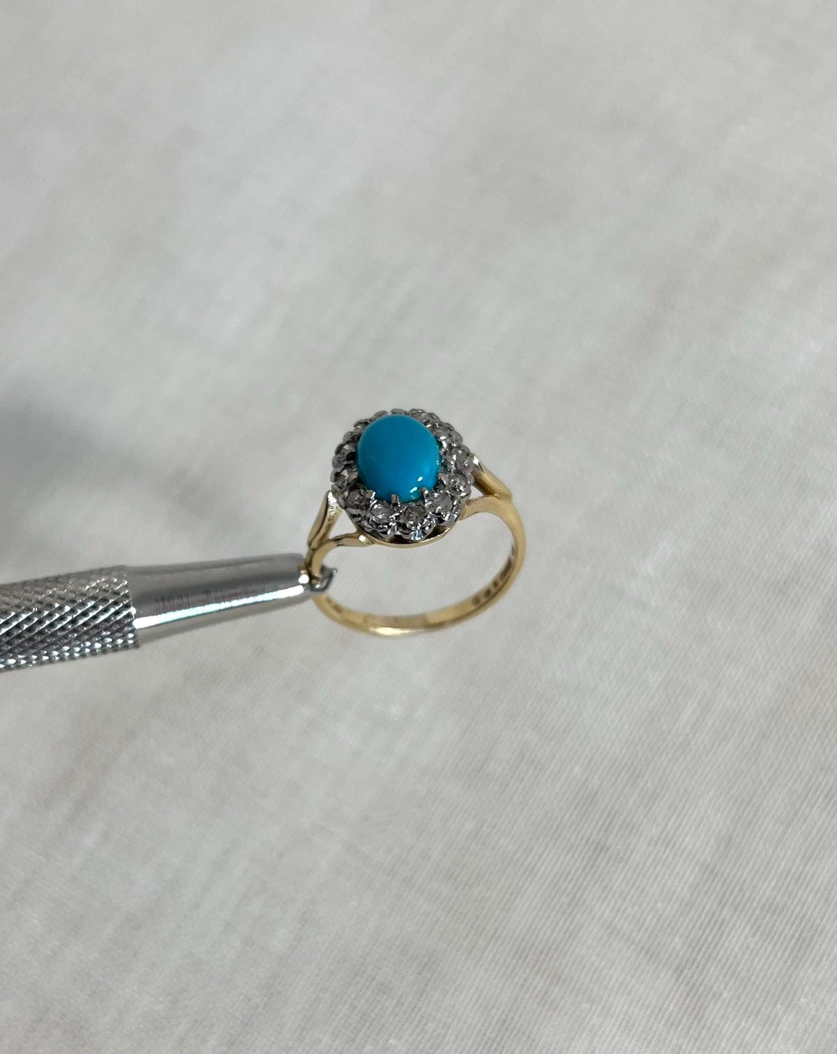 Gold ring with a turquoise gemstone and diamonds held by tweezers on a light fabric background