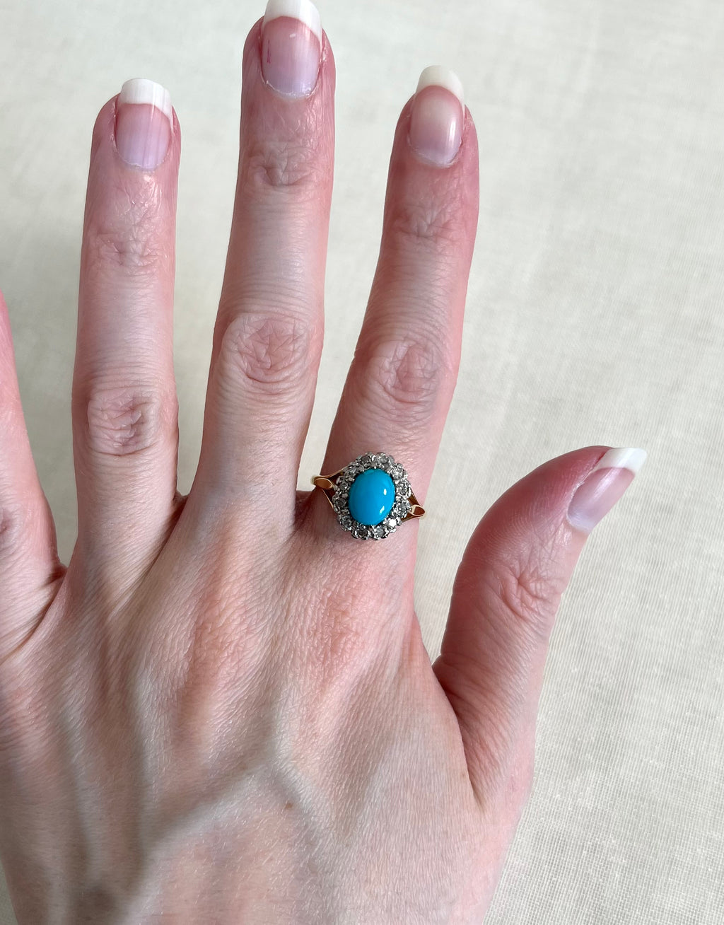 A close-up of a hand wearing a vintage-style ring with a round turquoise cabochon in a gold halo setting with diamonds, against a neutral beige fabric background.
