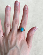 A close-up of a hand wearing a vintage-style ring with a round turquoise cabochon in a gold halo setting with diamonds, against a neutral beige fabric background.
